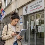 Žena ispred Nacionalne službe za zapošljavanje Zvezdara proverava informacije na telefonu i drži dokumenta pre odlaska u lokalni biro.